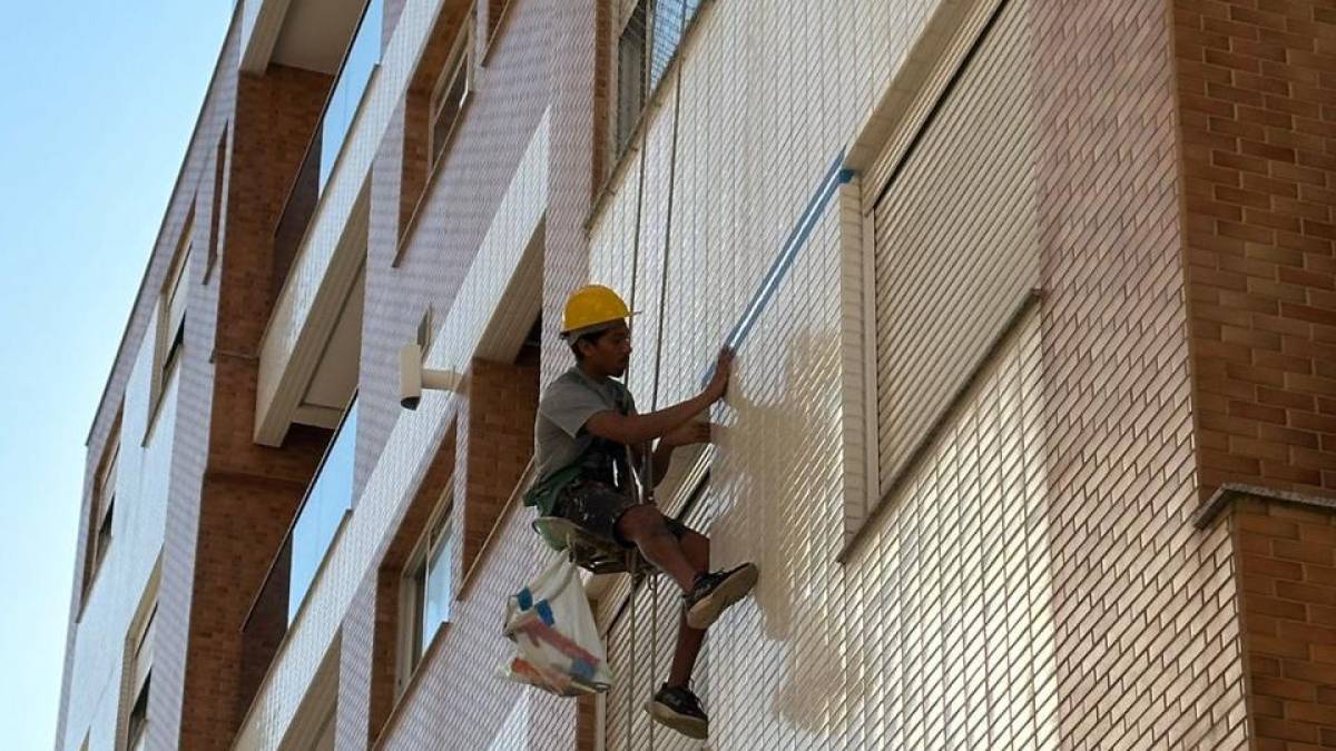 Profissional suspenso por corda lavando a fachada de um edifício, com a faixa já limpa visível ao lado da lança